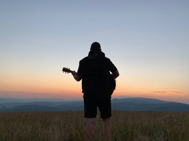 山の頂上にギターを持った男。夕日を背景にギターを弾く男のシルエット。山に楽器を持った男が. - 写真・画像