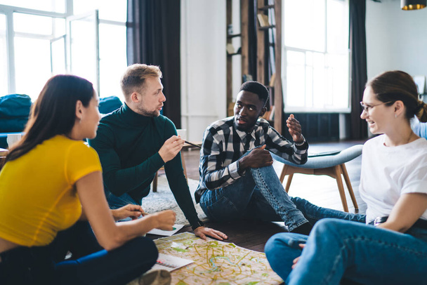 Group of content multiracial students in casual clothes gathering on floor with map and discussing trip details in light spacious apartment - Photo, image