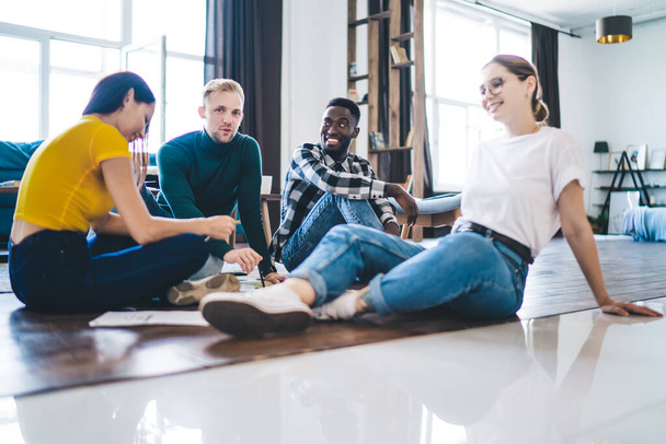 Full body positive young multiracial friends wearing casual outfits gathering on floor and having conversation while spending time together in spacious light living room - Foto, afbeelding