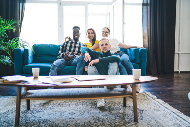 Content young multiracial coworkers in jeans gathering together while sitting on comfortable couch in front of table with cups and papers and looking at camera at home - Photo, Image