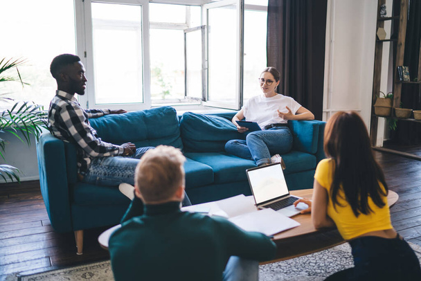Unrecognizable multiethnic partners with netbook speaking with colleagues on comfortable sofa near window while looking at each other in house - Valokuva, kuva