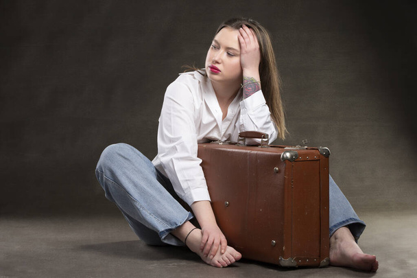 Beautiful girl sits with a vintage suitcase on a gray background. - Фото, изображение