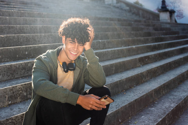 Portrait smiling young man man sitting on steps outside with mobile phone and headphones  - Photo, Image