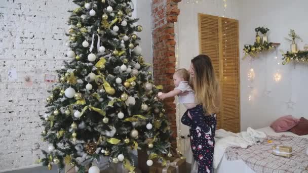Une jeune mère avec un petit fils dans ses bras décore un sapin de Noël à la maison - Séquence, vidéo