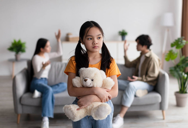 Lonely sad chinese teen girl with toy suffers from scandal of parents, young asian lady and guy quarreling at home - Foto, Imagem