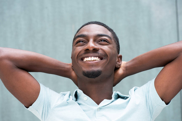 Κοντινό πλάνο πορτρέτο χαρούμενος νεαρός Αφροαμερικανός χαμογελά με τα χέρια πίσω από το κεφάλι σε μπλε φόντο και κοιτάζοντας προς τα πάνω - Φωτογραφία, εικόνα