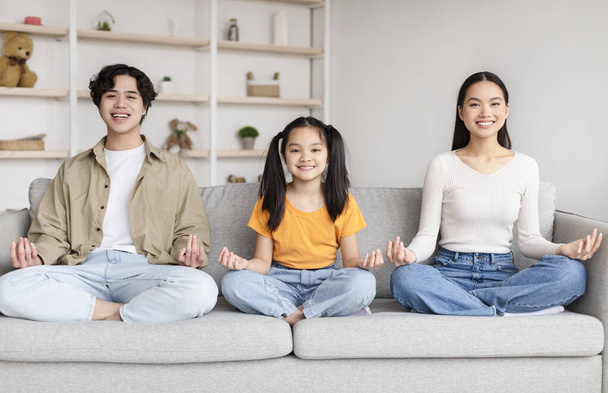 Smiling asian family sits in lotus position, practice yoga on sofa in minimalist living room interior at free time - Фото, изображение