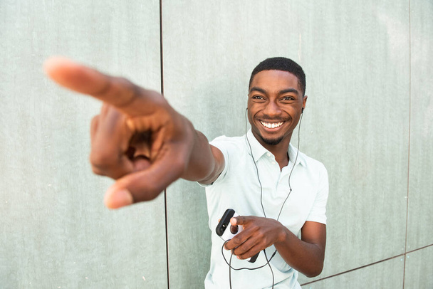 Close up portrait smiling young African American man listening to music with earbuds and mobile phone pointing finger - Photo, Image