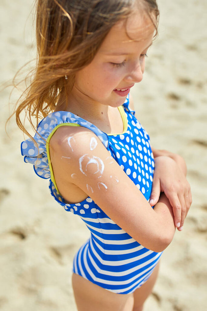 Kleines Mädchen mit Sonnencreme auf der Schulter in Form der Sonne am Strand stehend, Sonnencreme schützende Haut. Sommerferien. - Foto, Bild