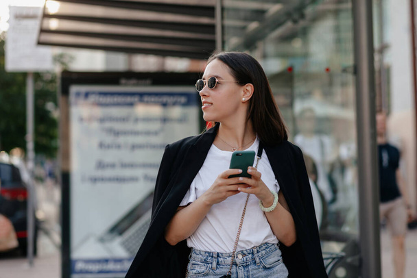 離れて見て、スマートフォンを使用してクローズアップスタイリッシュな女性。黒のジャケットと白のTシャツのサングラスのプリティーガールモデル。都市生活の概念 - 写真・画像