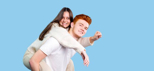 Two people brunette young woman and red hair young man making fun holding his girlfriend on the back over blue background, copy space. - Photo, image