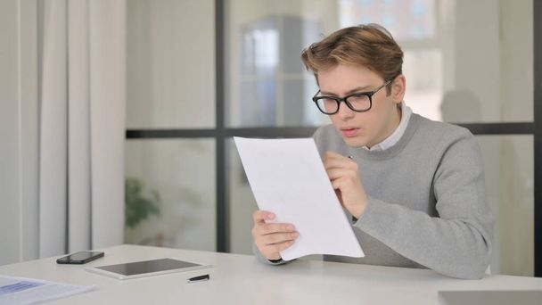Mladý muž čte dokumenty, zatímco sedí v moderní kanceláři - Fotografie, Obrázek