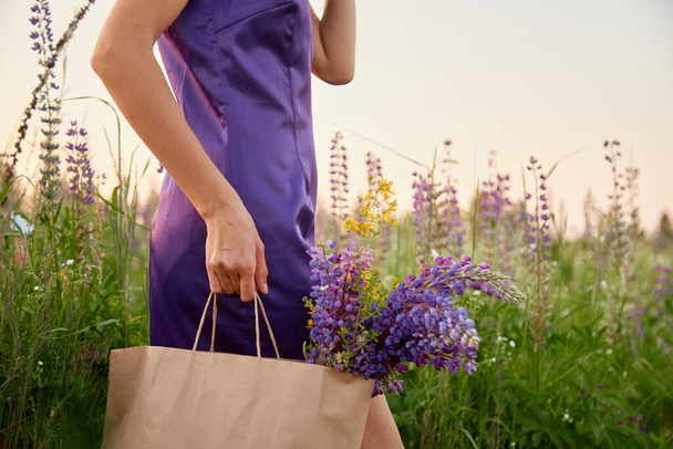 Woman in purple dress with craft bag of lupinus in the meadow. Lupin, lupine field with purple flowers at sunset. Natural products shopping concept. Wellness and natural concept. - Fotó, kép