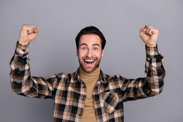 Photo of funny lucky guy dressed checkered shirt smiling rising fists isolated grey color background - Foto, Bild