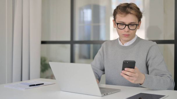 Mladý muž používající chytrý telefon při používání notebooku v moderní kanceláři  - Fotografie, Obrázek