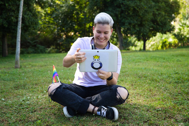 небинарный человек, сидящий в парке с табличкой с небинарным символом и флагом гей-гордости на коленях. Разнообразие и гей-прайд. Концепция недвоичной видимости. - Фото, изображение