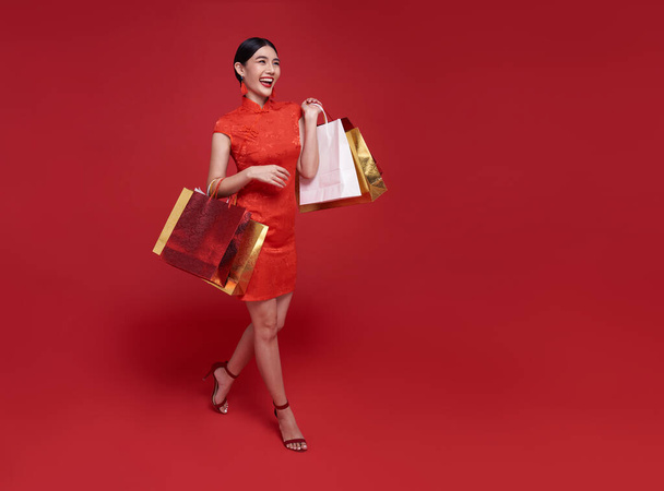 Mujer adicta a las compras asiáticas feliz usando cheongsam qipao vestido tradicional sosteniendo bolsa aislada sobre fondo rojo. Feliz año nuevo chino - Foto, Imagen