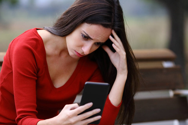 Worried woman sitting on a bench checking smart phone content in a park - Photo, image