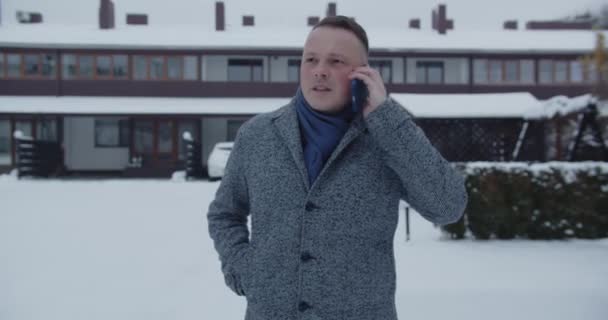The man is focused on talking on a mobile phone and walks down a snowy street - Materiał filmowy, wideo