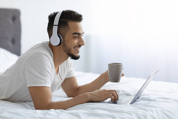 Χιλιετής Άραβας με ακουστικά που ξεκουράζονται στο κρεβάτι με laptop και καφέ - Φωτογραφία, εικόνα