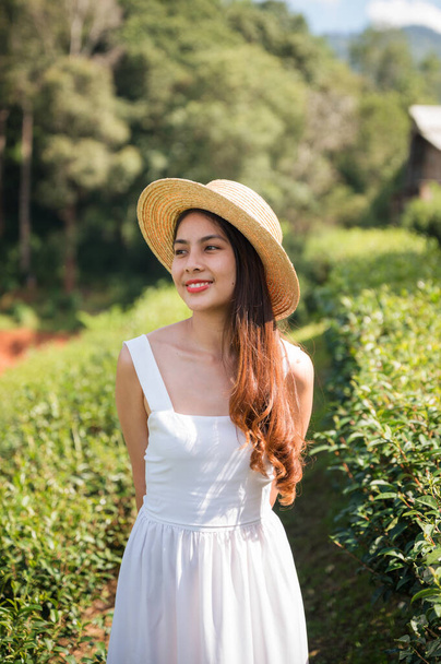 Hermosa mujer asiática usando sombrero en vestido blanco de pie en plantación de té en día soleado - Foto, imagen