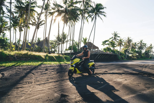Side view full length male biker in summer wear and yellow helmet riding motorcycle on black sandy path along tropical palm in sunny day - Фото, изображение