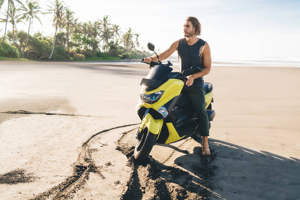Full body of confident young bearded male traveler riding modern motorbike on sandy beach between waving ocean and palm trees on sunny day - Foto, Imagen