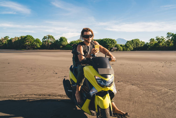Full length of brutal young trendy guy in casual outfit and goggles chilling on sandy beach sitting on yellow motorbike and text messaging on mobile phone on sunny day - 写真・画像
