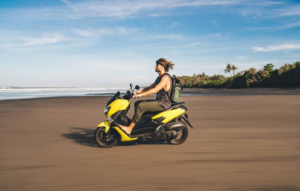 Side view of full length male biker driving electric scooter among sandy beach while looking away and spending summer in tropical country - Фото, зображення