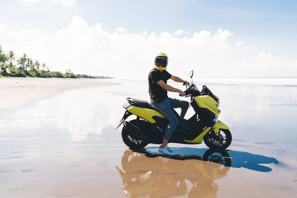 Full body side view of anonymous young male biker in trendy outfit and helmet riding yellow scooter on wet sandy coast near waving ocean on sunny day - Fotografie, Obrázek