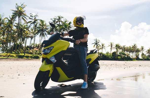 From below of young male biker in casual wear and helmet on motorcycle enjoying beauty of nature while standing against tropical background with palm trees and cloudy sky - Photo, Image