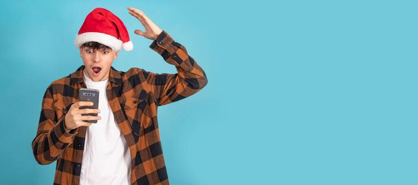 erstaunt Teenager Junge mit Weihnachtsmann Hut Blick auf Handy isoliert auf blauem Hintergrund - Foto, Bild