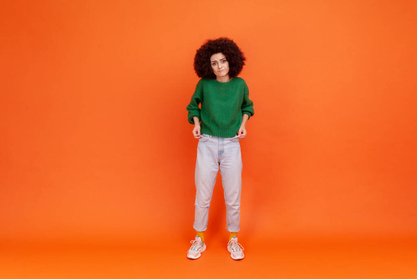 Full length portrait of woman with Afro hairstyle wearing green casual style sweater showing empty pockets, has no money, jobless female. Indoor studio shot isolated on orange background. - Fotó, kép