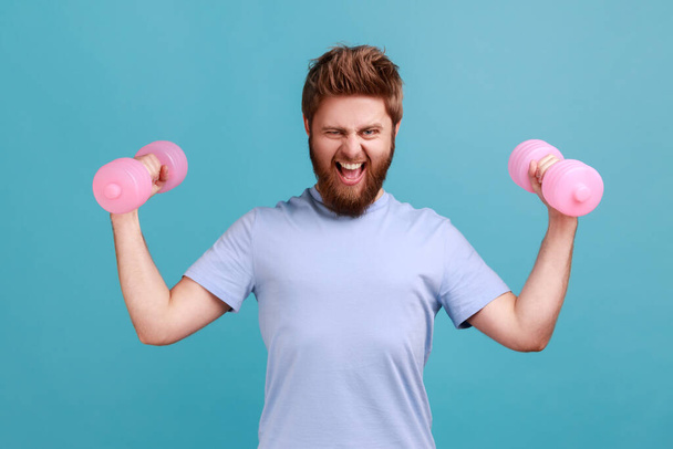 Portrait of bearded man holding pink dumbbells in hands and looking at camera with an effort, pumping up muscles, healthy lifestyle. Indoor studio shot isolated on blue background. - Photo, Image