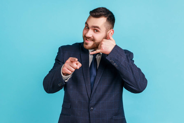 You contact us! Portrait of positive man with beard in dark suit making gesture with fingers dial my number or call me back and pointing at you. Indoor studio shot isolated on blue background. - Photo, image