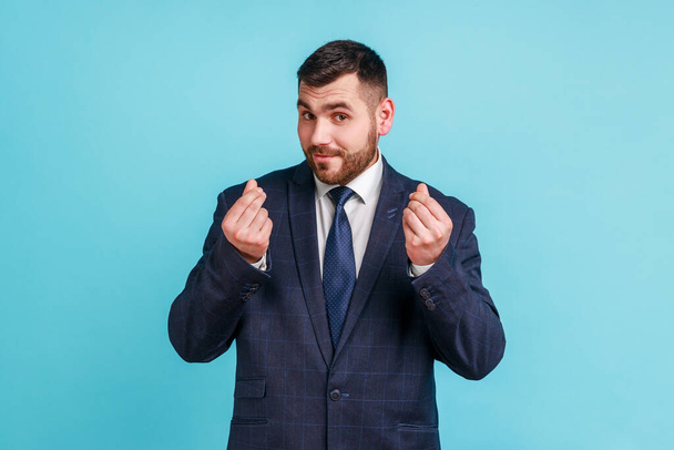 Portrait of handsome bearded young man wearing official style suit standing and looking at camera with money or italian gesture with hand. Indoor studio shot isolated on blue background. - Fotoğraf, Görsel