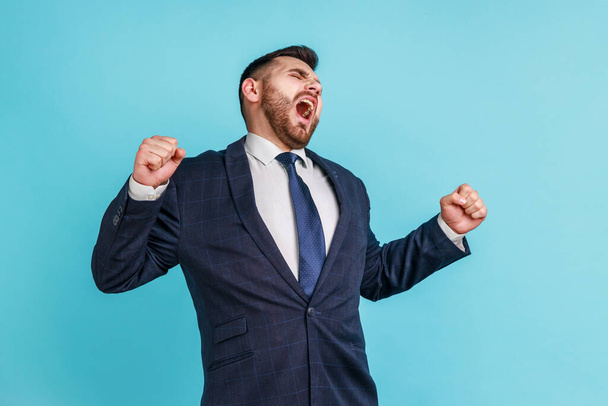 Young attractive businessman with beard wearing dark official style suit yawning and raising hands up, feeling fatigued of overtime work Indoor studio shot isolated on blue background. - Foto, afbeelding