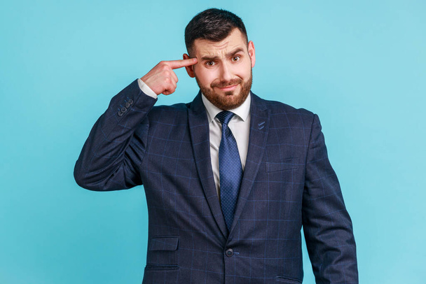 Crazy idea! Portrait of man in dark suit showing stupid gesture, looking at camera with condemnation and blaming for insane plan, dumb suggestion. Indoor studio shot isolated on blue background. - Photo, Image