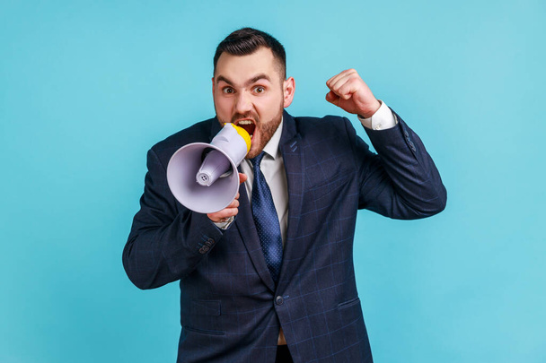 Angry serious businessman wearing dark official style suit loudly speaking screaming holding megaphone, announcing important message. Indoor studio shot isolated on blue background. - Fotoğraf, Görsel