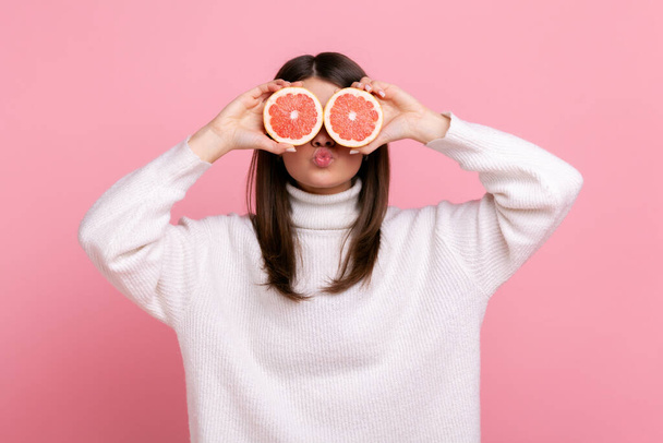 Attractive girl covering eyes with half slices of grapefruit, keeps lips pout, sending air kisses, wearing white casual style sweater. Indoor studio shot isolated on pink background. - Foto, afbeelding