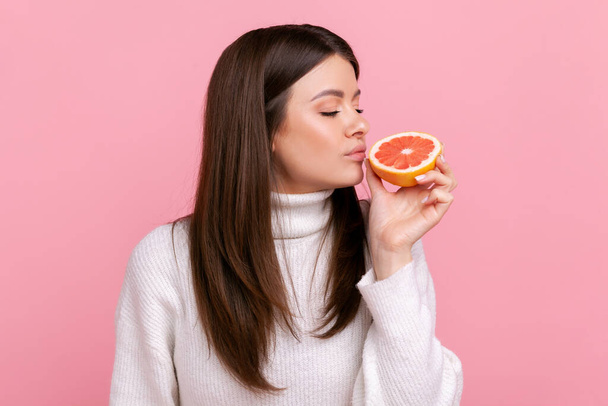 Beautiful female smelling half slice grapefruit, enjoying tasty vitamin fruits, healthy nutrition, wearing white casual style sweater. Indoor studio shot isolated on pink background. - 写真・画像