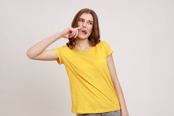 Funny comical teenager girl with brown hair picking finger to nose and showing tongue, looking at away, having fun, fooling around, bad manners. Indoor studio shot isolated on gray background. - 写真・画像