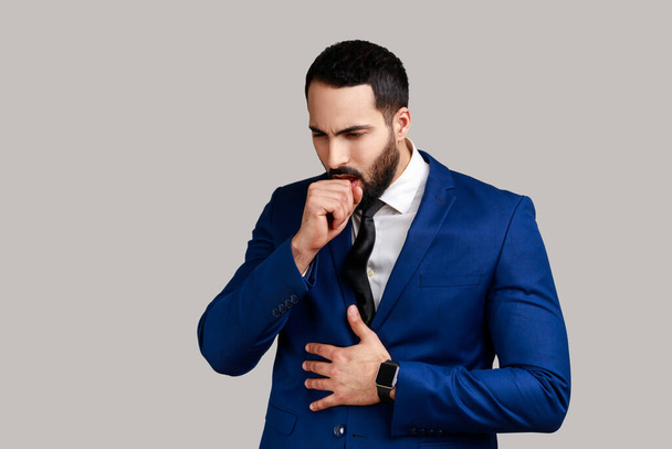 Portrait of unhealthy bearded man coughing, catches cold, having high temperature, having influenza symptom, wearing official style suit. Indoor studio shot isolated on gray background. - Foto, immagini