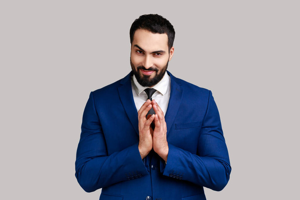 Cunning bearded man clasping hands and planning evil tricky prank or scheming, cheating with sly smile, wearing official style suit. Indoor studio shot isolated on gray background. - Fotoğraf, Görsel