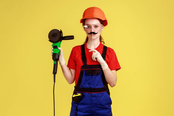 Portrait of funny worker woman standing grinding saw and mustache on stick, looking at camera, wearing overalls and protective helmet. Indoor studio shot isolated on yellow background. - Foto, afbeelding