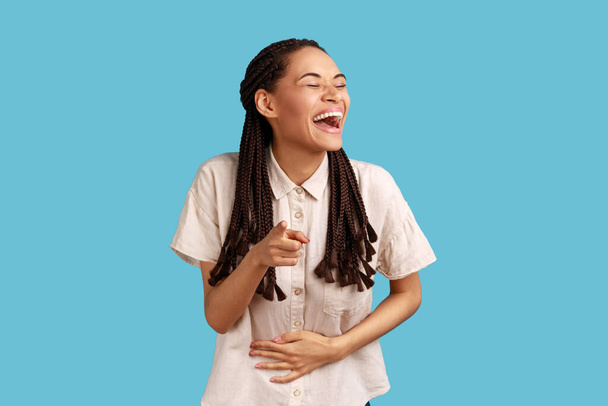 Pleased woman with black dreadlocks pointing finger at camera, gestures we need you, blaming someone, has funny reaction, wearing white shirt. Indoor studio shot isolated on blue background. - Fotografie, Obrázek