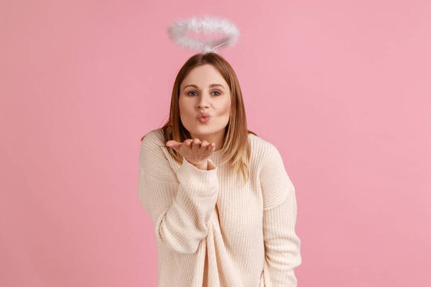 Portrait of romantic charming blond angelic woman with nimbus over head looking at camera and sending air kisses, wearing white sweater. Indoor studio shot isolated on pink background. - 写真・画像