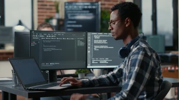 Retrato de desarrollador afroamericano usando portátil para escribir código sentado en el escritorio con algoritmo de análisis de múltiples pantallas - Imágenes, Vídeo
