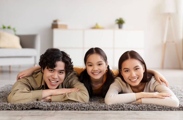 Smiling happy chinese millennial lady, guy and teen daughter resting on floor in living room interior, look at camera - Foto, Imagem
