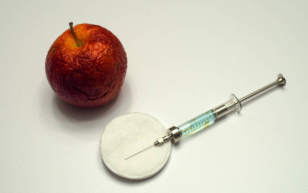Ένα ζαρωμένο μήλο και μια γυάλινη σύριγγα σε ένα λευκό απομονωμένο στέλεχος. Θεραπεία για τα γηρατειά. Ένα αναζωογονητικό εμβόλιο. - Φωτογραφία, εικόνα